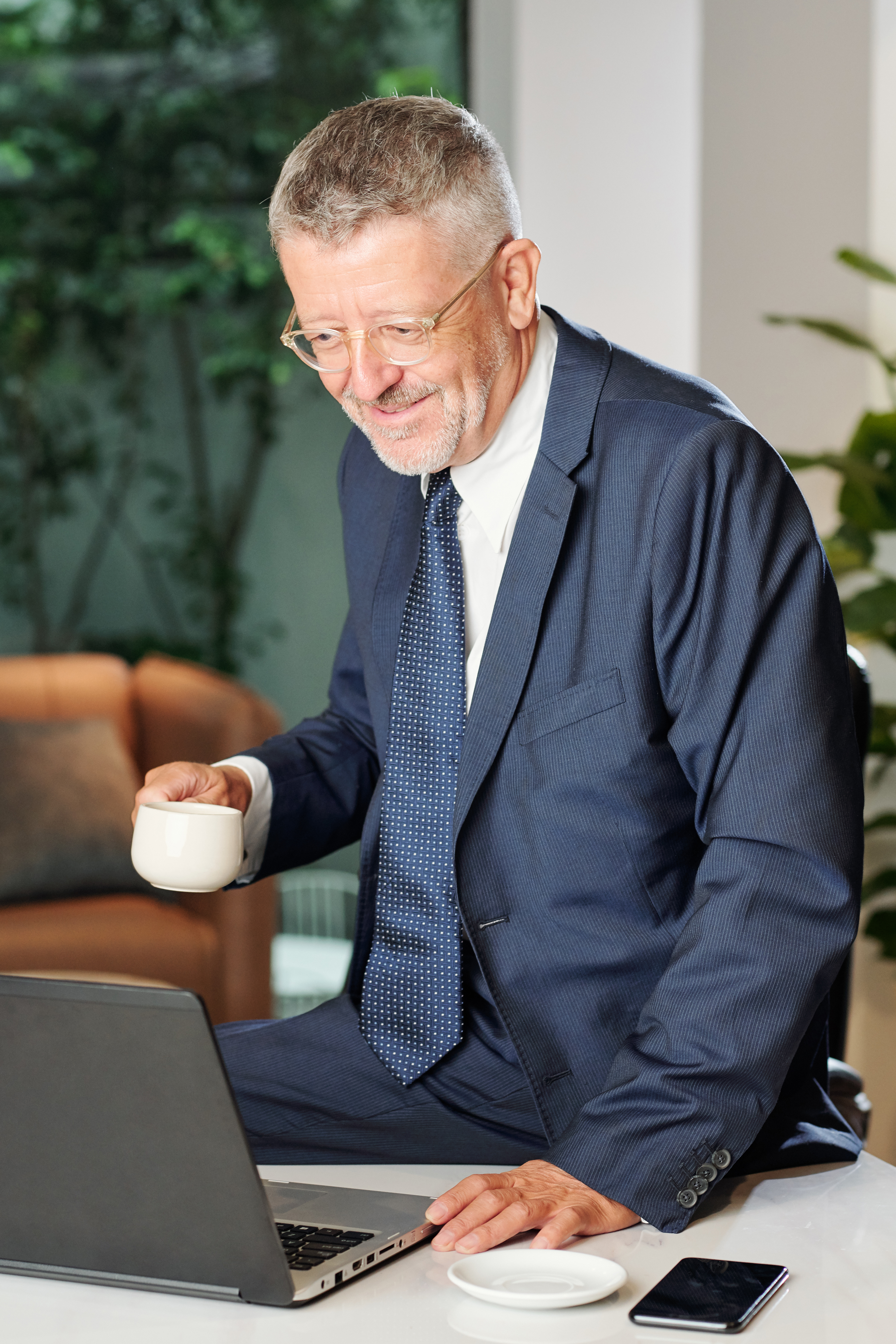 Smiling aged businessman drinking morning coffee and reading e-mails on laptop screen