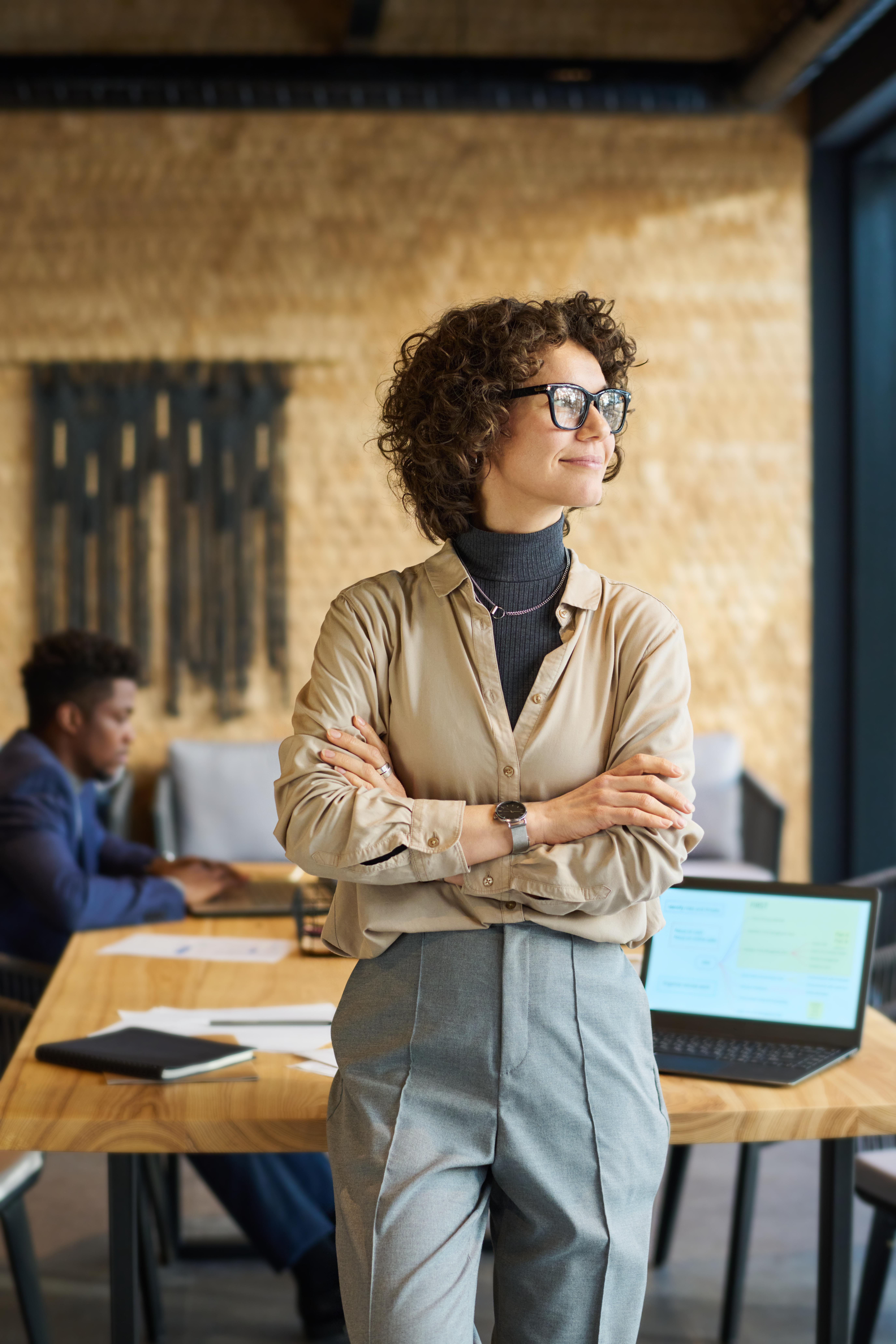 Young pretty business leader in quiet luxury attire looking through window while standing in front of camera by workplace against coworker
